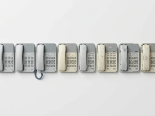 Rij gelijkende gereviseerde kantoortelefoons op wit oppervlak, variërend van onberispelijk tot licht versleten, in minimalistische compositie.