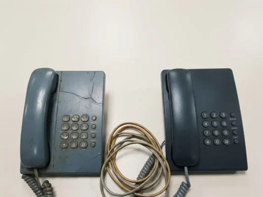 Verouderde kantoortelefoon met gebarsten behuizing naast een gerenoveerd vervangend toestel op een licht oppervlak.