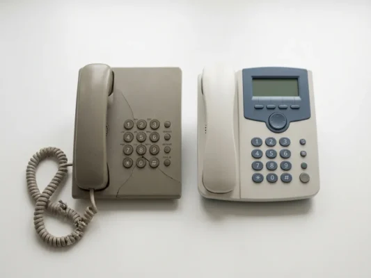 Verouderde kantoortelefoon met gebarsten behuizing en rafelige kabel naast een nieuw vervangend toestel op wit oppervlak.