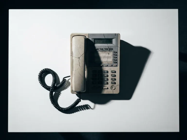 Verouderde kantoortelefoon met haarscheurtjes en losse kabel op wit oppervlak, zijdelings belicht met scherpe schaduwen.